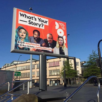University signage promoting 'what's your story' fir #sheffdocfest. Sufnafe is at a university campus with a red background and famous comedians on the sign.